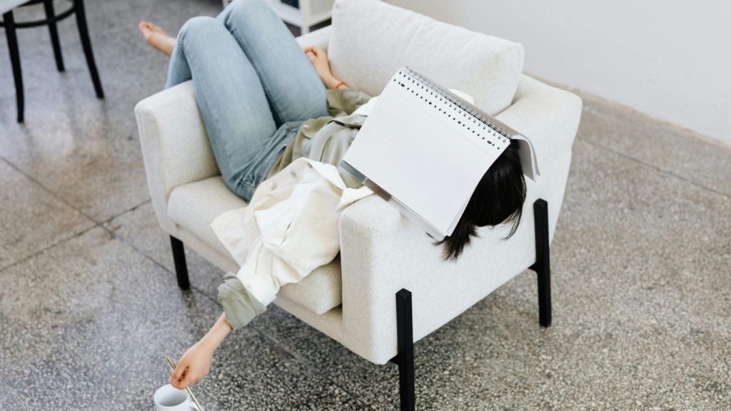 Woman lying on an armchair with a book on face, holding a coffee. Casual and relaxing atmosphere.