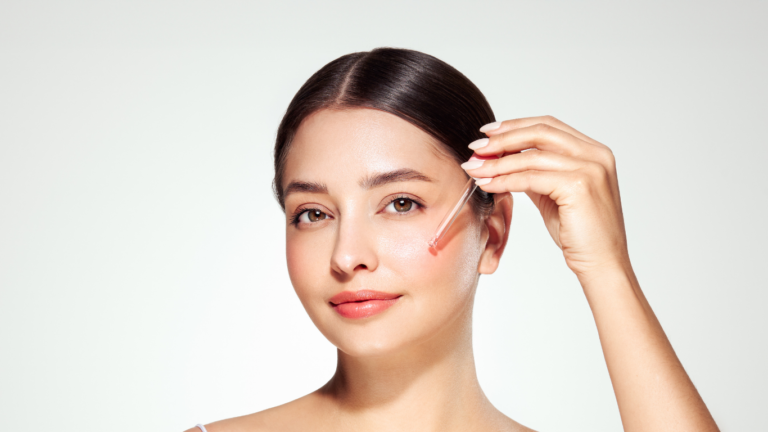 A young woman with smooth skin applies a peach-colored facial serum to her cheek with a dropper while looking at the camera against a light gray background.
