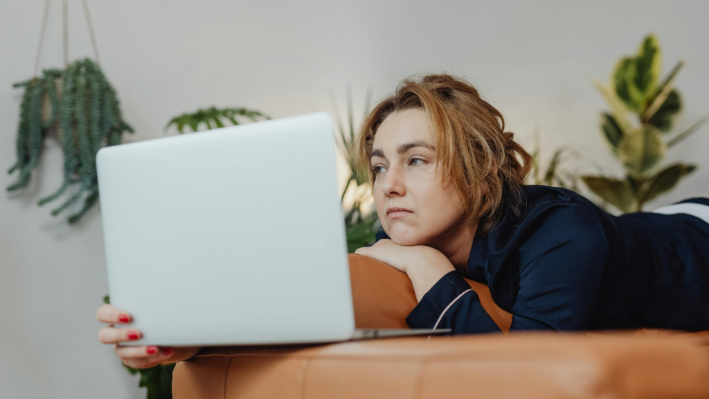 Woman lying on an sofa looking to her Macbook lazly holding a coffee. She doesn't seem motivated