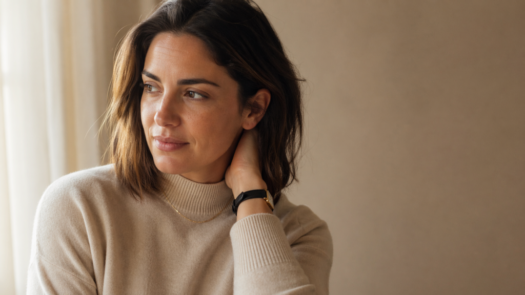 woman in her mid-thirties in a soft cream sweater and gold jewelry, photographed in warm natural light in a quiet editorial style, her gaze thoughtful and turned slightly away from the camera